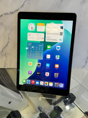 A black Apple iPad tablet standing upright on a clear display stand, showing the home screen with colorful widgets and app icons against a marble-patterned background.