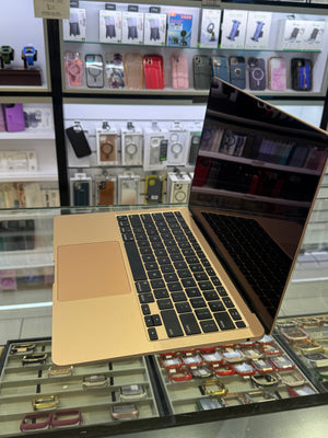 A gold Apple MacBook laptop with a black screen and keyboard is displayed on a glass counter inside a retail store, with watch and phone accessories visible in the background.