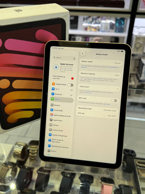 An iPad displaying the Apple Account battery health settings screen, standing upright in a retail store display with Apple products in the background.