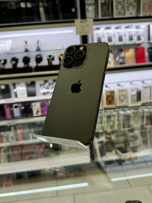 A dark gray Apple iPhone Pro shown from the back, resting on a clear acrylic display stand inside a retail store with blurred shelves of accessories in the background.