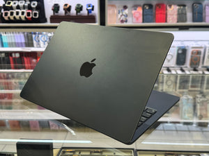 A black Apple MacBook laptop shown open at an angle, resting on a glass display counter inside a retail store. The Apple logo is clearly visible on the back of the laptop lid.