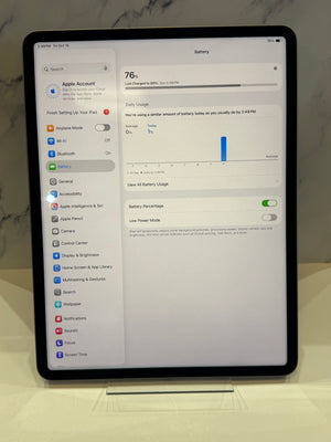 An Apple iPad standing upright on a small clear display stand, showing the Settings app screen with the Battery tab open. The tablet is photographed indoors against a light marble-like surface and wall.