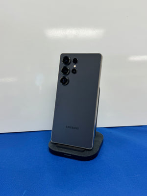 A Samsung smartphone shown from the back, standing upright on a small black charging stand against a white wall with a blue surface below.