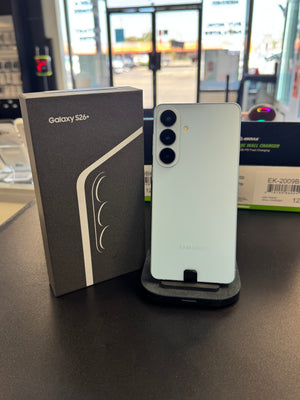 A Samsung Galaxy S24+ smartphone in a pale mint green color stands on a small black stand next to its original retail box on a dark tabletop, with a blurred store background.
