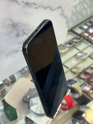 A black smartphone with a glossy screen resting on a white marble-patterned surface, with a blurred watch display case in the background.