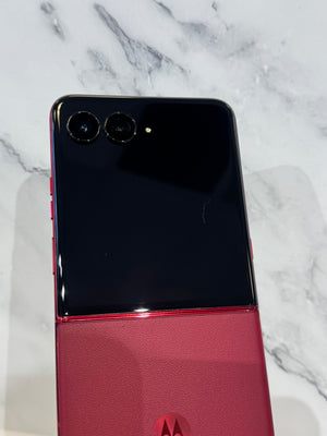 A Motorola flip-style smartphone shown closed from the back, with a glossy black top half and a textured pink-red bottom half, photographed on a white marble surface.