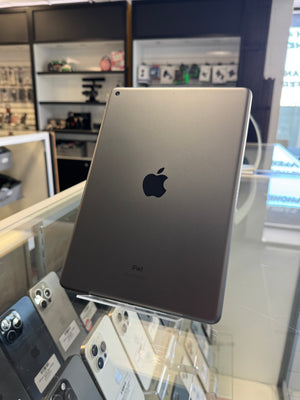 An Apple iPad shown from the back, standing on a display stand inside a retail store. The device has a dark gray metallic finish with the iconic black Apple logo in the center.