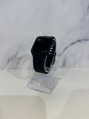 A black smartwatch with a rectangular screen and black band, displayed on a clear acrylic stand against a white marble-patterned background.