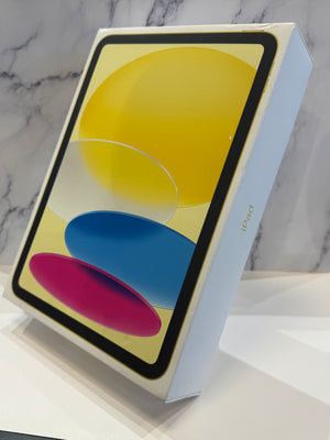An Apple iPad in its original white box, displaying a colorful screen graphic with yellow, blue, and pink abstract shapes, set against a light marble-patterned background.