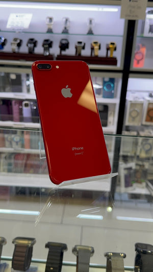 A red Apple iPhone 7 Plus shown from the back, resting on a clear acrylic display stand inside a retail store. The Apple logo is visible in the center, with watch displays in the background.