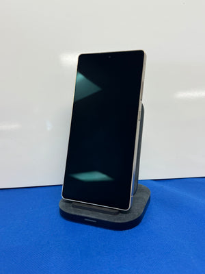 A black smartphone with a large screen, shown standing upright on a dark gray charging dock or stand against a white background with a blue surface below.