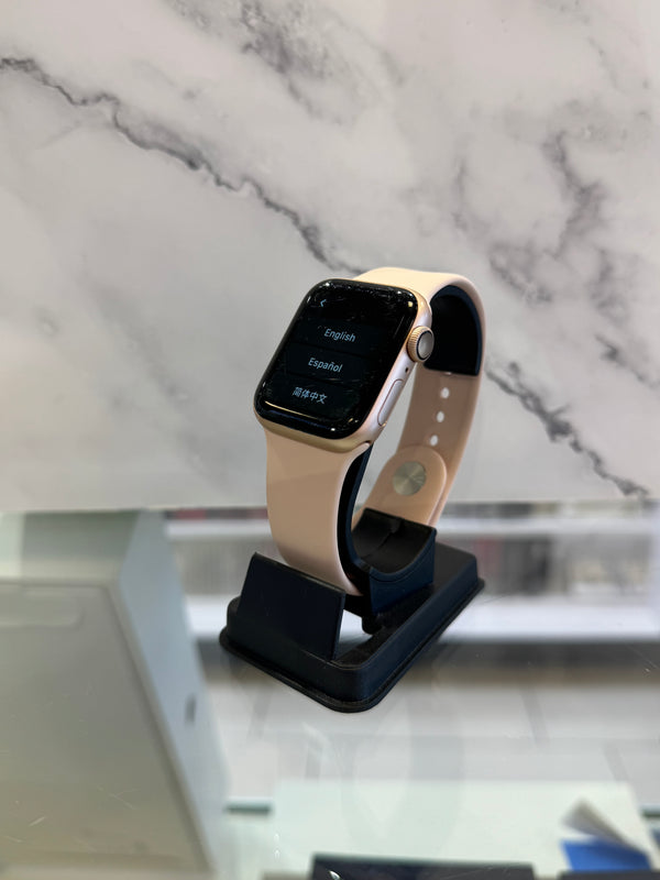 A smartwatch with a light beige band and a gold-toned case, displayed on a black stand against a white marble-patterned background.