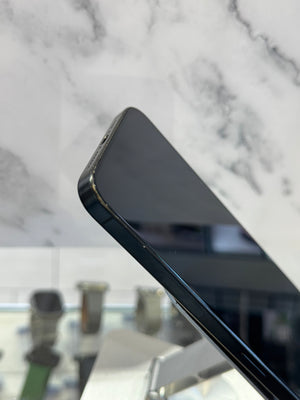 Close-up of a black smartphone corner resting on a white marble surface, with a blurred indoor background visible through the glass.