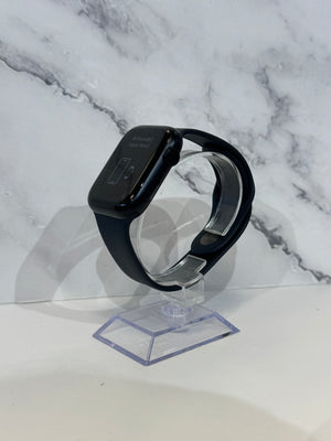 A black smartwatch with a rectangular face and black band, displayed on a clear acrylic stand against a white marble-patterned background.