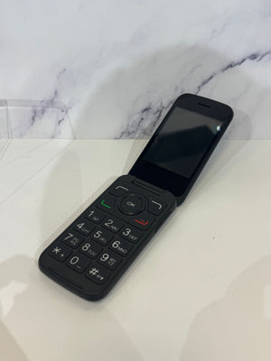 An open flip phone resting on a white tabletop showing a dark keypad and a dark screen. 
