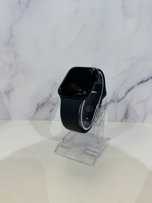 A black smartwatch with a rectangular glossy screen and a dark band, displayed on a clear acrylic stand against a white marble-patterned background.