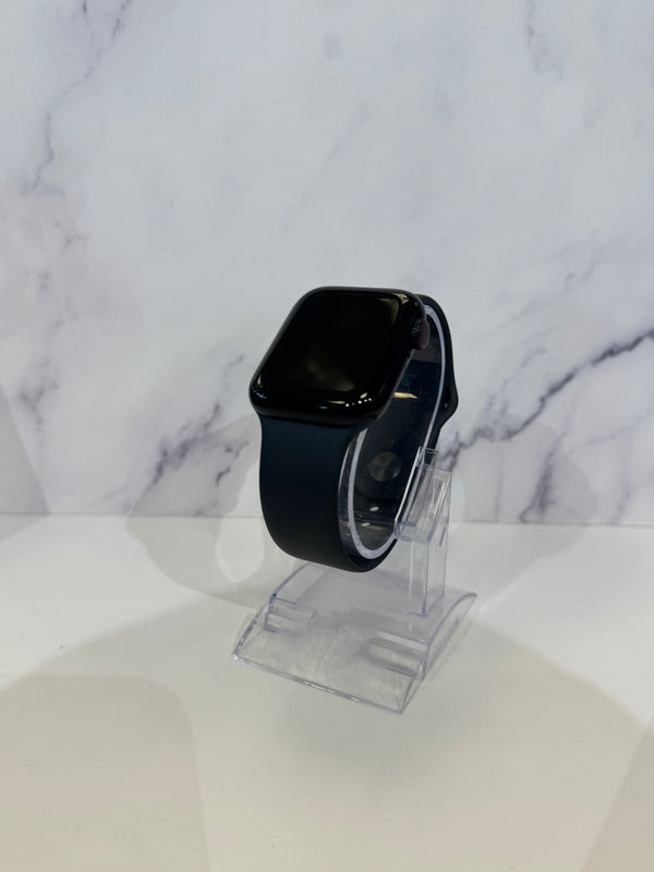 A black smartwatch with a rectangular glossy screen and a dark band, displayed on a clear acrylic stand against a white marble-patterned background.