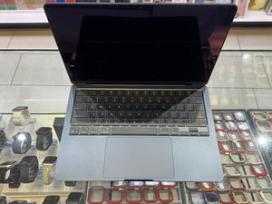 A silver aluminum laptop with a black screen and keyboard, shown open on a glass display counter in a retail store. Watch and accessory displays are visible in the background.