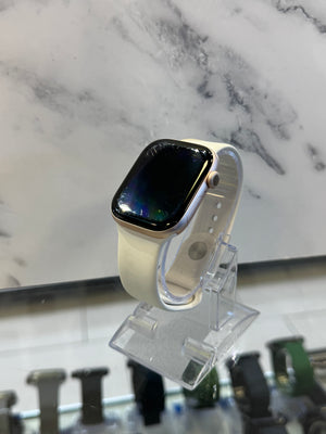 A smartwatch with a light beige band and a gold-toned case, displayed on a clear acrylic stand against a white and gray marble-patterned background.
