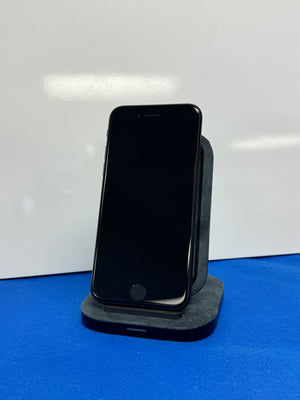 A black smartphone with a blank screen stands upright on a dark gray charging dock against a white background with a blue surface below.