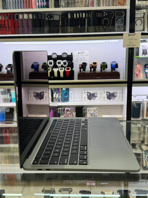 A silver Apple MacBook laptop is displayed on a glass counter inside a retail store, with shelves of various smartwatches and phone cases visible in the background.