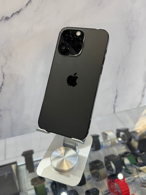 A black Apple iPhone 12 Pro shown from the back, resting on a silver metal phone stand in a retail display with marble-patterned wall and other watches in the background.