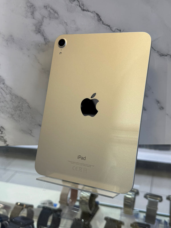 A gold-colored iPad shown from the back, standing upright on a clear display stand against a white marble-patterned wall.