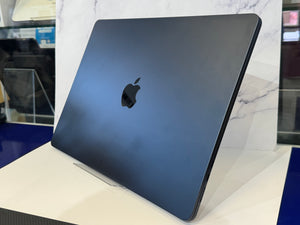 A closed Apple laptop in a dark metallic blue finish, standing upright on a white display stand in front of a marble-patterned backdrop.