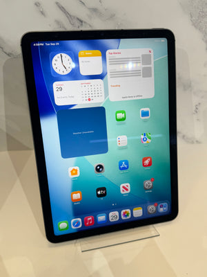 An iPad with a black bezel displaying its home screen, resting on a clear acrylic stand against a light marble-patterned background.