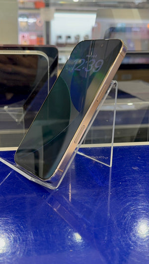 A smartphone with a dark screen and a metallic rose gold or light gold frame, displayed on a clear acrylic stand on a blue reflective surface.