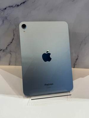 A silver Apple iPad mini with a blue-to-silver gradient back cover, standing upright on a clear display stand against a white surface and a gray marble-patterned background.
