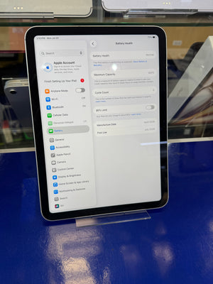 A tablet device shown from above on a glossy blue display surface, with its screen turned on and displaying settings pages including Apple Account and battery health.