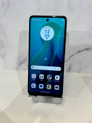 A black Google Pixel smartphone with its screen on, displaying the home screen, resting on a clear plastic display stand against a white marble-patterned background.
