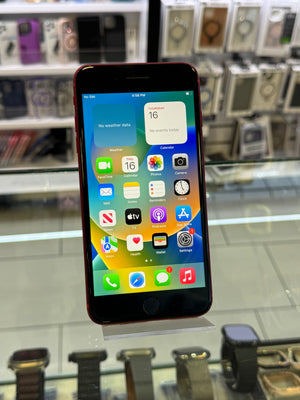 A red iPhone with a black front bezel and colorful app icons on its screen, displayed on a clear stand inside a retail store with watch straps and boxed electronics in the blurred background.