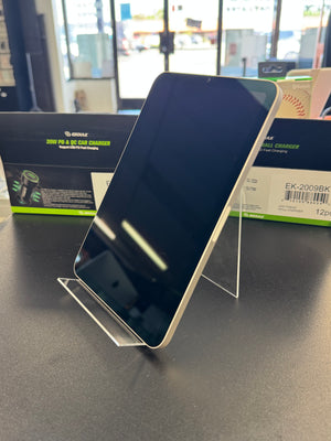 A black smartphone displayed upright on a clear acrylic stand on a dark tabletop, with product packaging boxes visible in the background.