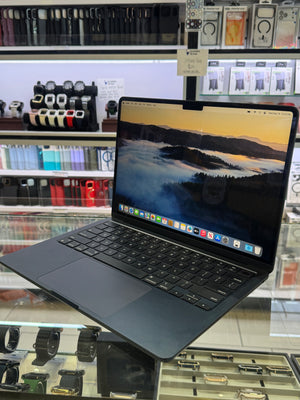 A dark gray Apple MacBook laptop with an open screen displaying a mountain landscape wallpaper, set on a glass display counter inside a retail store with shelves of accessories in the background.