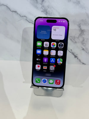 A black iPhone with a purple and white home screen wallpaper, resting on a small silver metal stand against a white marble-patterned background.