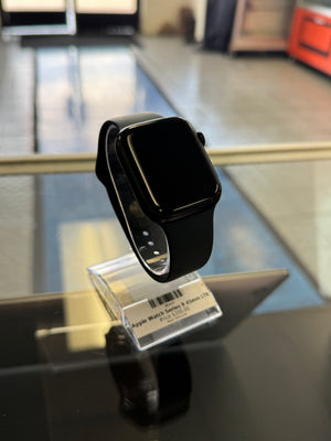 A black smartwatch with a black band displayed on a glass surface next to its retail packaging tag.