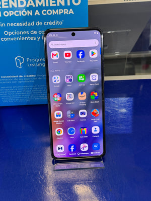 A black smartphone with a large front display showing various colorful app icons, resting on a blue surface next to a blue promotional box.