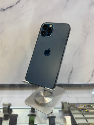 A dark blue-gray Apple iPhone 12 Pro shown from the back, resting on a silver magnetic display stand against a white marble-patterned background.