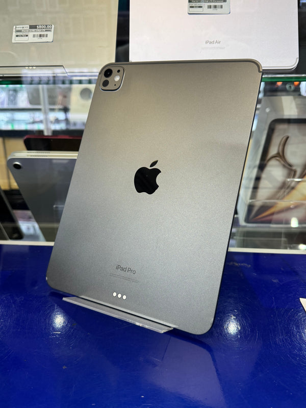Silver Apple iPad Pro back view with the Apple logo and camera module visible, photographed on a blue display surface with other devices partially visible around it.