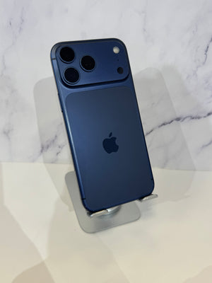 A dark blue Apple iPhone 12 Pro shown from the back, resting on a silver metal display stand against a white marble-patterned background.