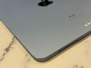 Close-up of the back corner of a silver Apple iPad resting on a light marble surface, showing the Apple logo, iPad text, and speaker grilles.