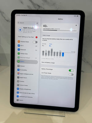 A black tablet, likely an iPad, standing upright on a clear display stand on a light marble surface. The screen is on and displaying the Apple Settings app with battery information.
