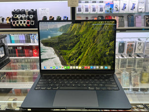 A dark gray Apple MacBook laptop with an open screen displaying a lush green coastal landscape wallpaper, set on a glass display counter inside a retail store.