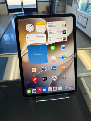 A black iPad Pro tablet on a clear display stand, showing the home screen with various app icons and widgets. The tablet is photographed in a retail or showroom environment with glass surfaces and blurred background elements.