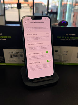 A smartphone on a charging stand displaying the battery health and charging settings screen, with product boxes in the blurred background.
