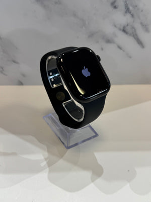 A black Apple Watch with a black sport band, displayed on a clear acrylic stand against a white marble-patterned background.
