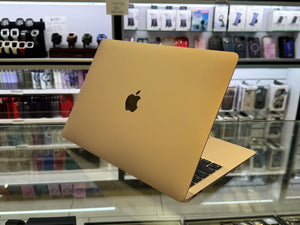 A gold Apple MacBook laptop is shown open on a glass display counter inside a retail store, with shelves of accessories and electronics blurred in the background.