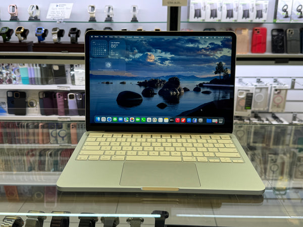 A silver Apple MacBook laptop with a cream-colored keyboard and a colorful ocean landscape wallpaper, displayed on a glass counter inside a retail store.